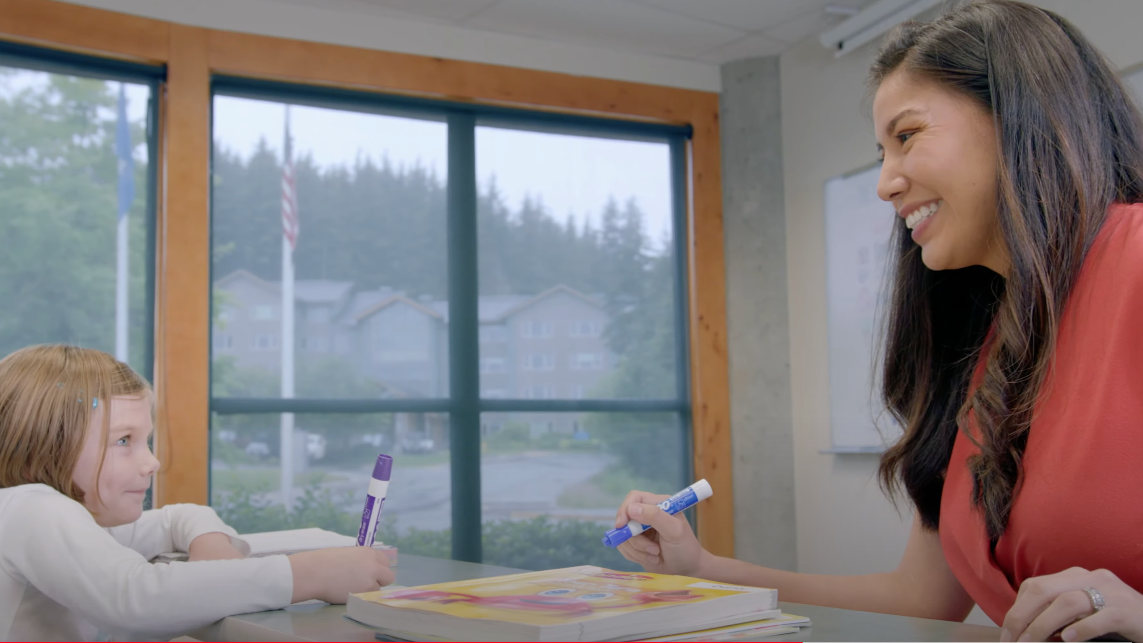 A UAS teacher smiles back at a young student as they write together.