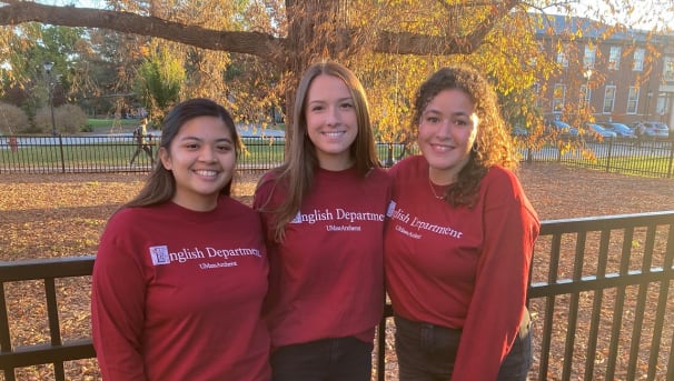 Three smiling English majors wear red English Department shirts as they pose near the South College.