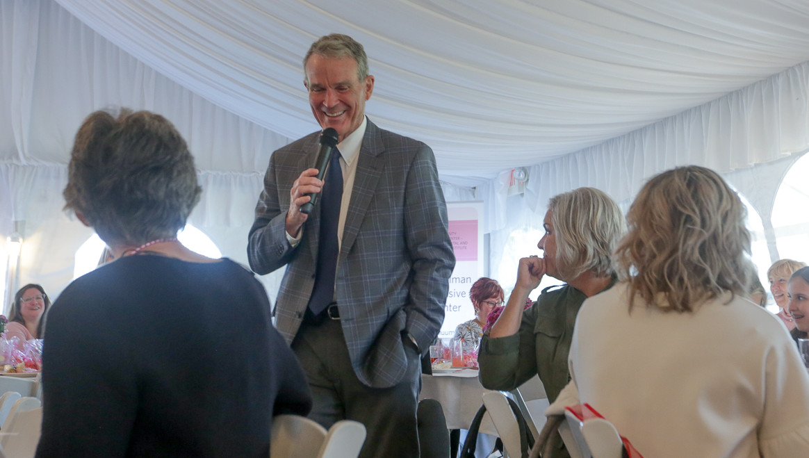 A man, holding a microphone, stands in the center of tables, speaking