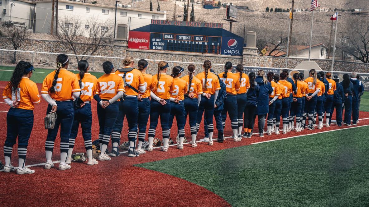 Softball team in uniform, orange shirts, standing in a line facing scoreboard