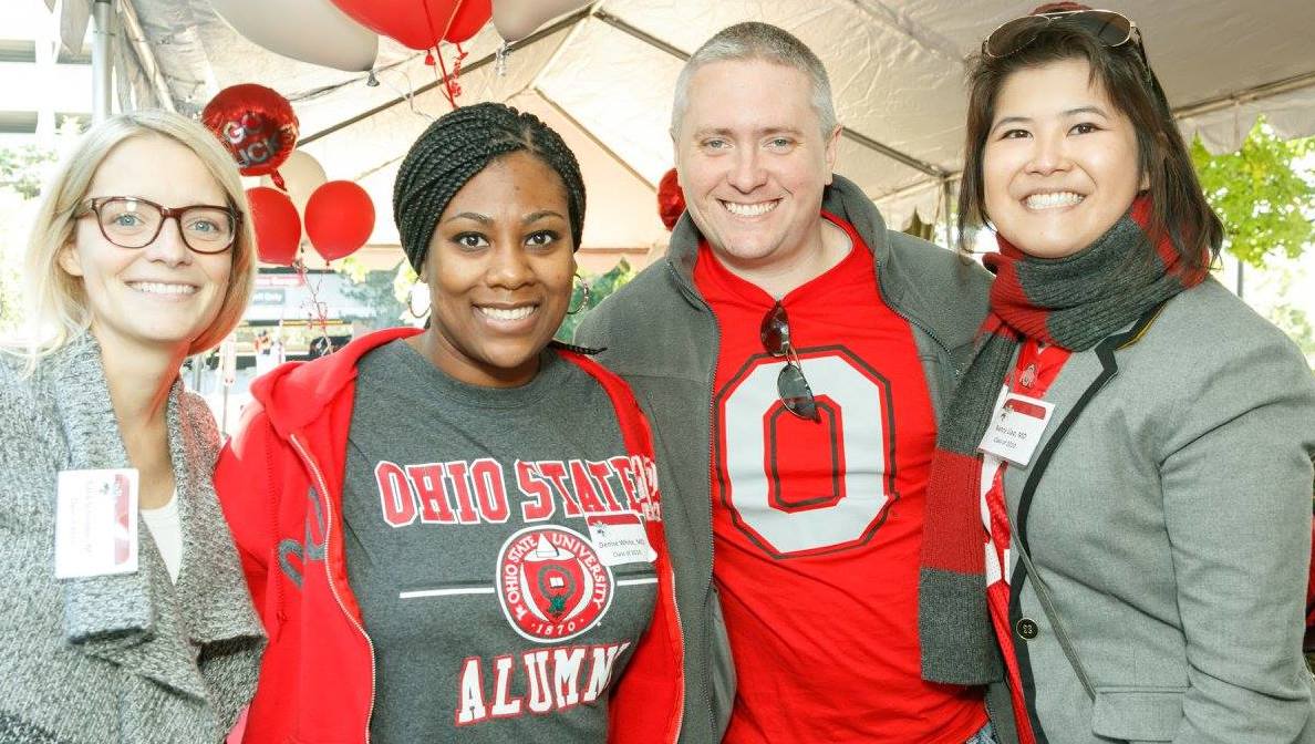 Four alums pose together beneath a large, white tend. All wear Ohio State gear