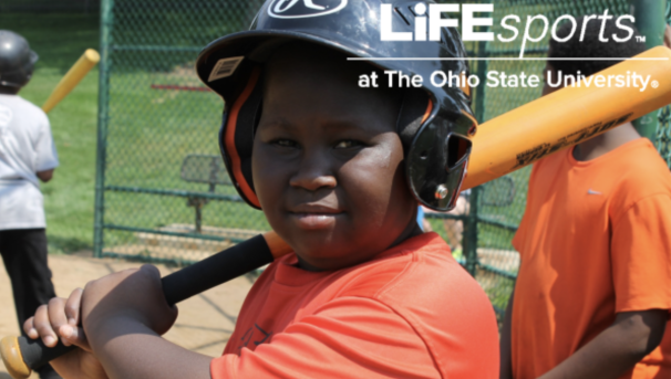 A boy with a baseball bat resting on his shoulder. Behind him is a tall, green fence and bench