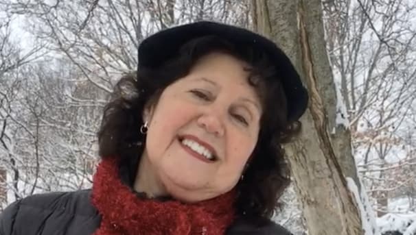 A woman wearing a red scarf and black hat smiling at the camera. She stands outside in the snow.