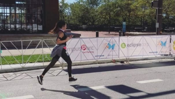 Kristine Pollard running in a marathon. She runs next to white fencing with various sponsor's logos