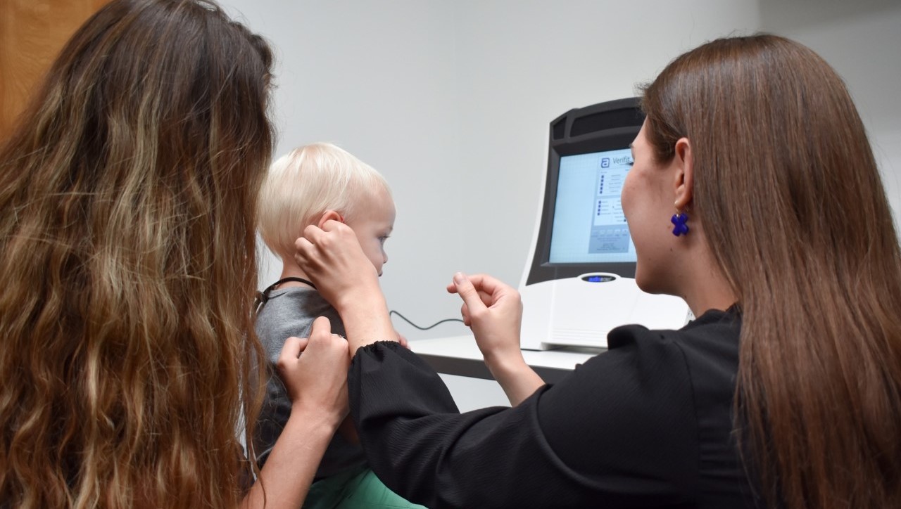 A nurse works on a child for their Hearing Aid Fitting. He sits on his mother's lap