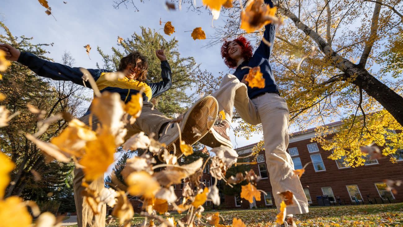 Students kicking up autumn leaves on campus during a fall afternoon