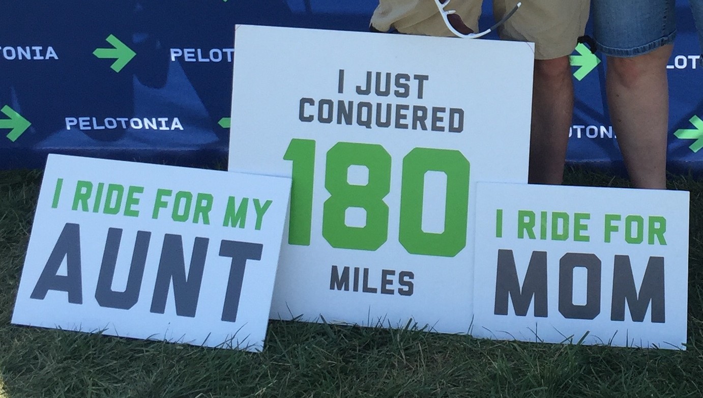 Three signs read 'I ride for my aunt', 'I just conquered 180 miles', and 'I ride for my mom'