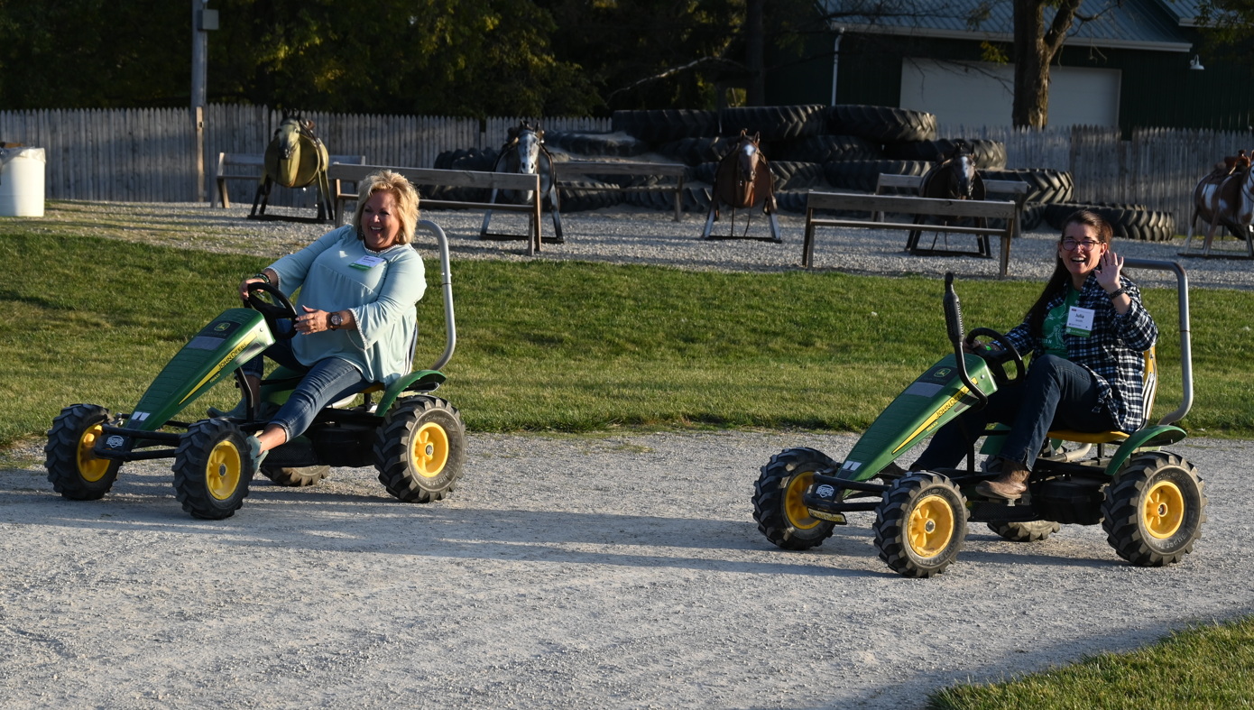 Two adult women are riding peddle cart tractors and laughing together. One is waving at the camera.