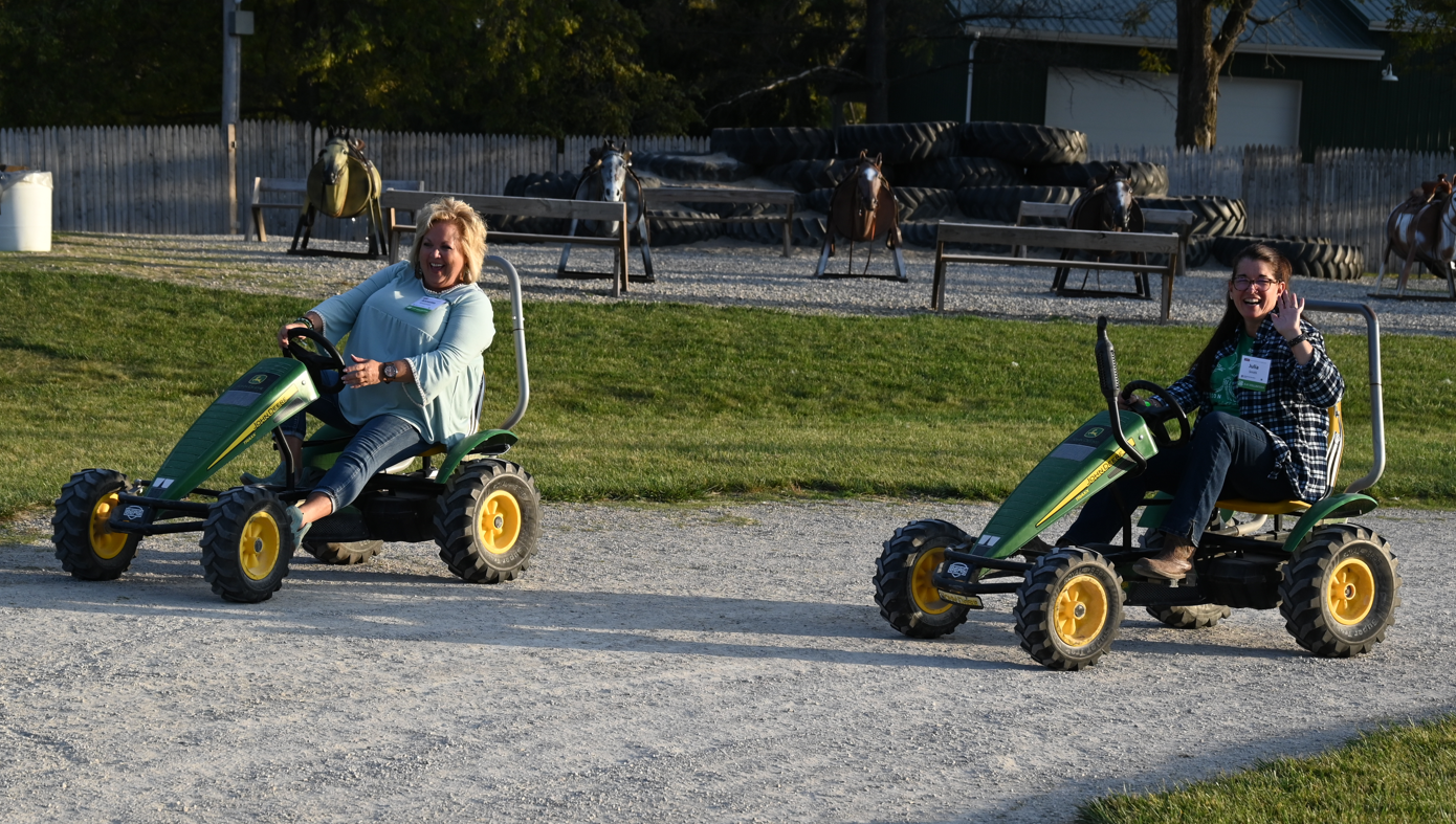 Two adult women are riding peddle cart tractors and laughing together. On is waving at the camera.