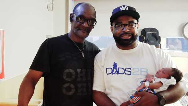 Two men stand side by side. The one wearing the Dads2B shirt holds his newborn