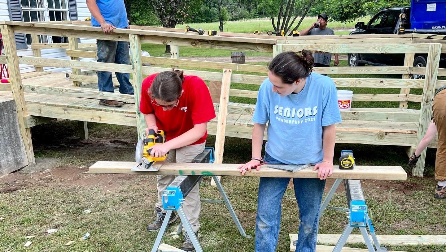Two individuals work on sawing at a piece of long plywood