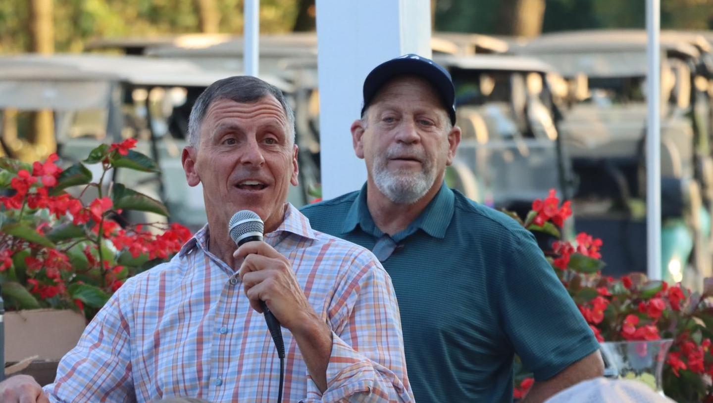 Man speaking into microphone with second man behind him, in front of a line of golf carts