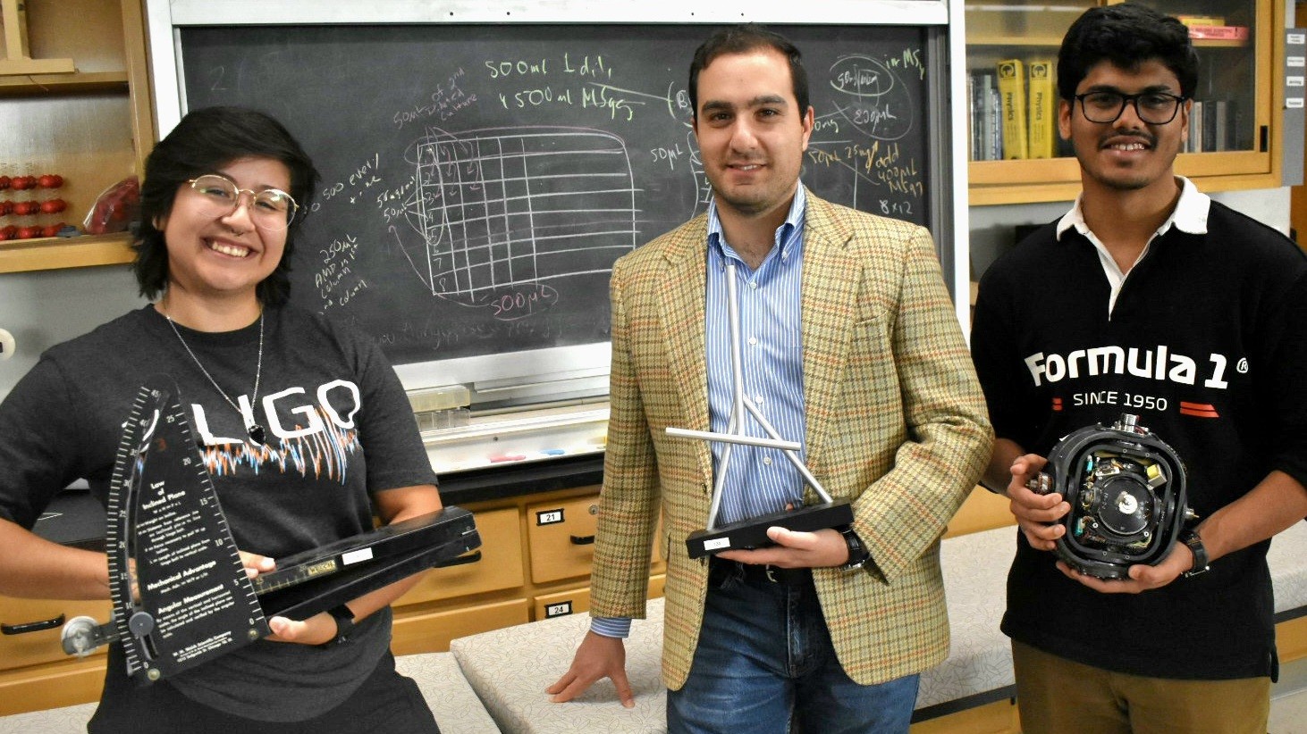 Three students smile and hold physics equipment in a lab