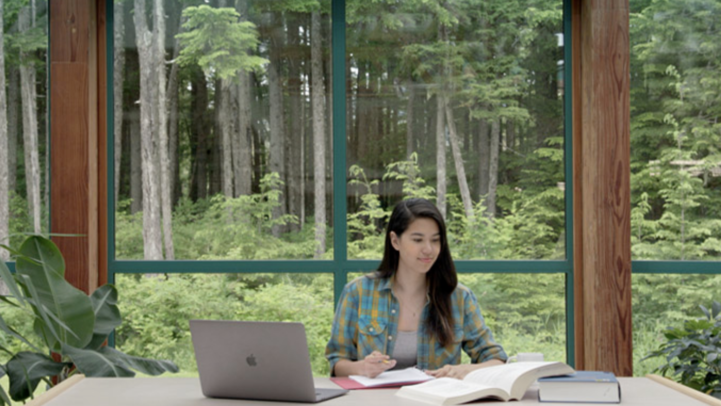 A student studies with open book and laptop in front of a window.