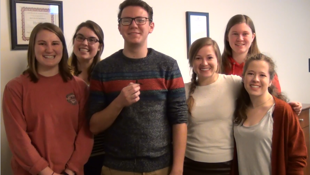 Six individuals smiling for a picture in front of a white wall with various photos
