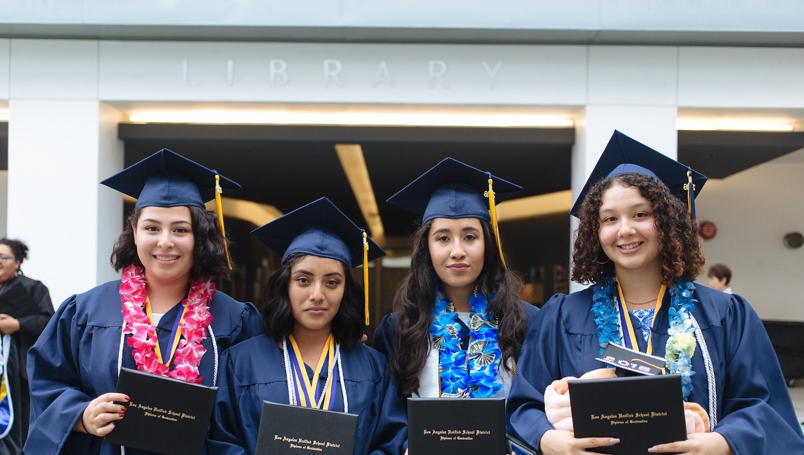 Four UCLA Community School graduates smiling in front of the library with their diplomas.