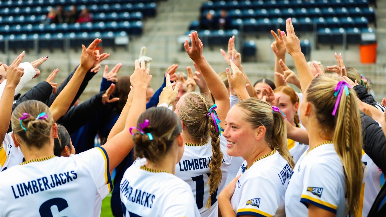 The NAU Women's Soccer team stands in a huddle with their hands up. One player smiling.