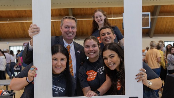 Six people in gray and orange attire smiling through a white picture frame.