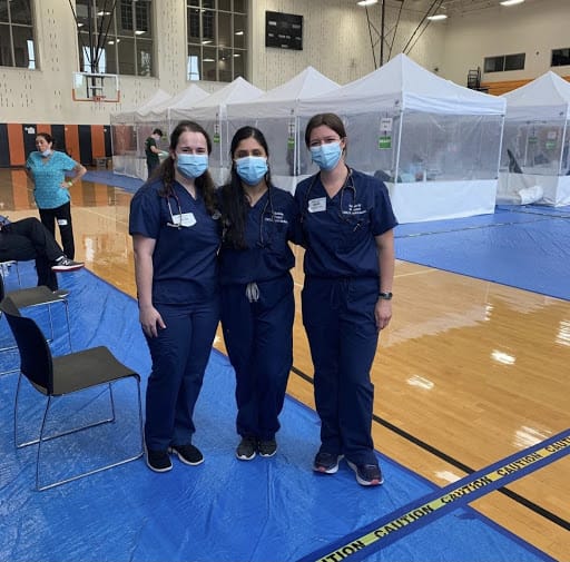 Three masked women in scrubs stand in school gym