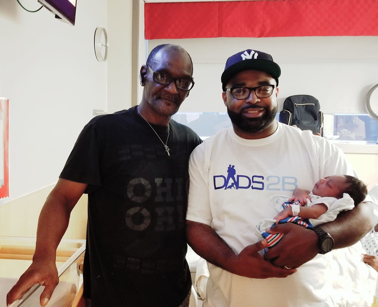 Two men stand side by side, one holding a baby