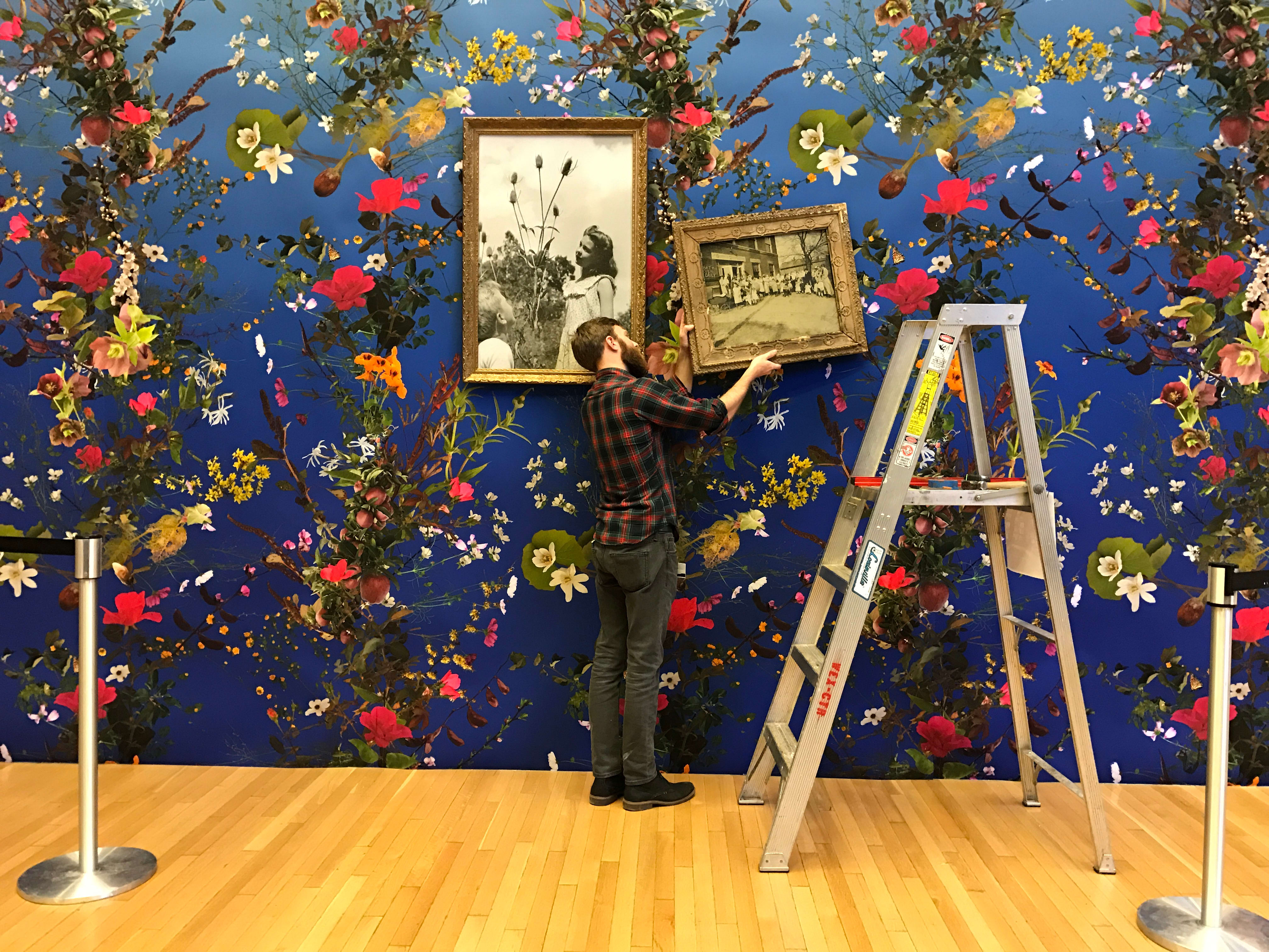 A man hangs two frames against a floral wallpaper