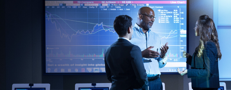 2 students speaking to a professor in front of a screen showing stock market trends