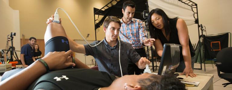 Two professors standing over a student testing another students knee 