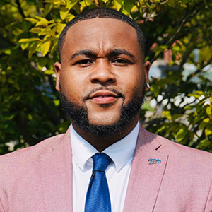 Demondre Peak's professional headshot. There is a tree with bright green leaves behind him