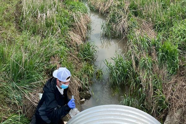 A student crouched in front of a sewer gate. They are standing on the side of where the water had pooled on the muddy ground