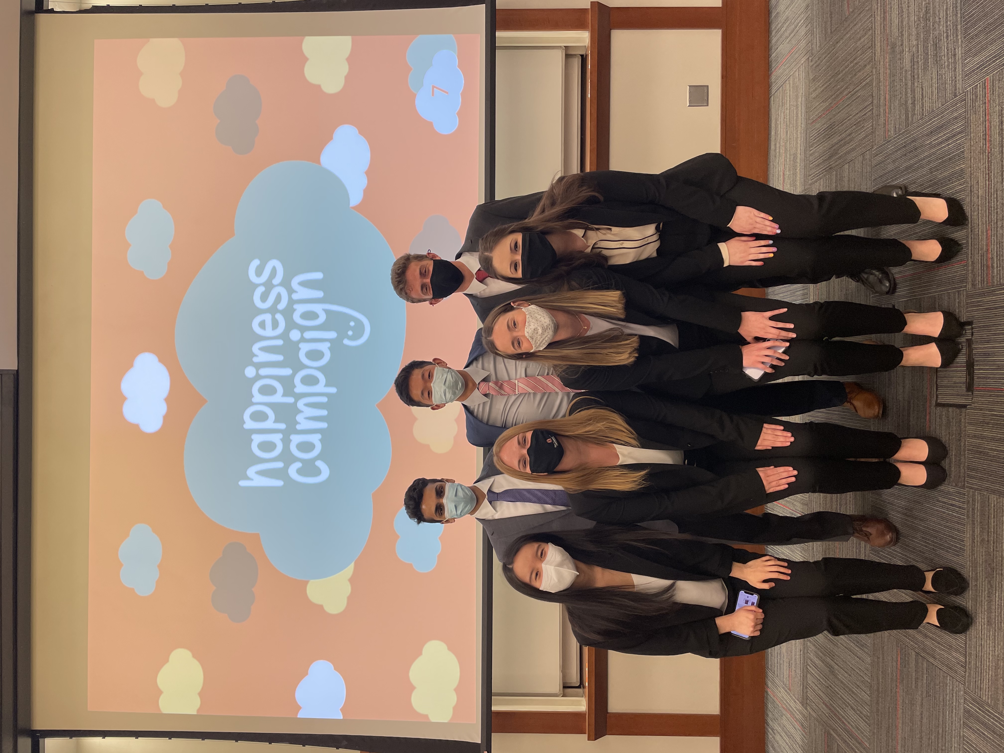 Seven individuals, all wearing masks, posed before a 'happiness campaign' projection. 