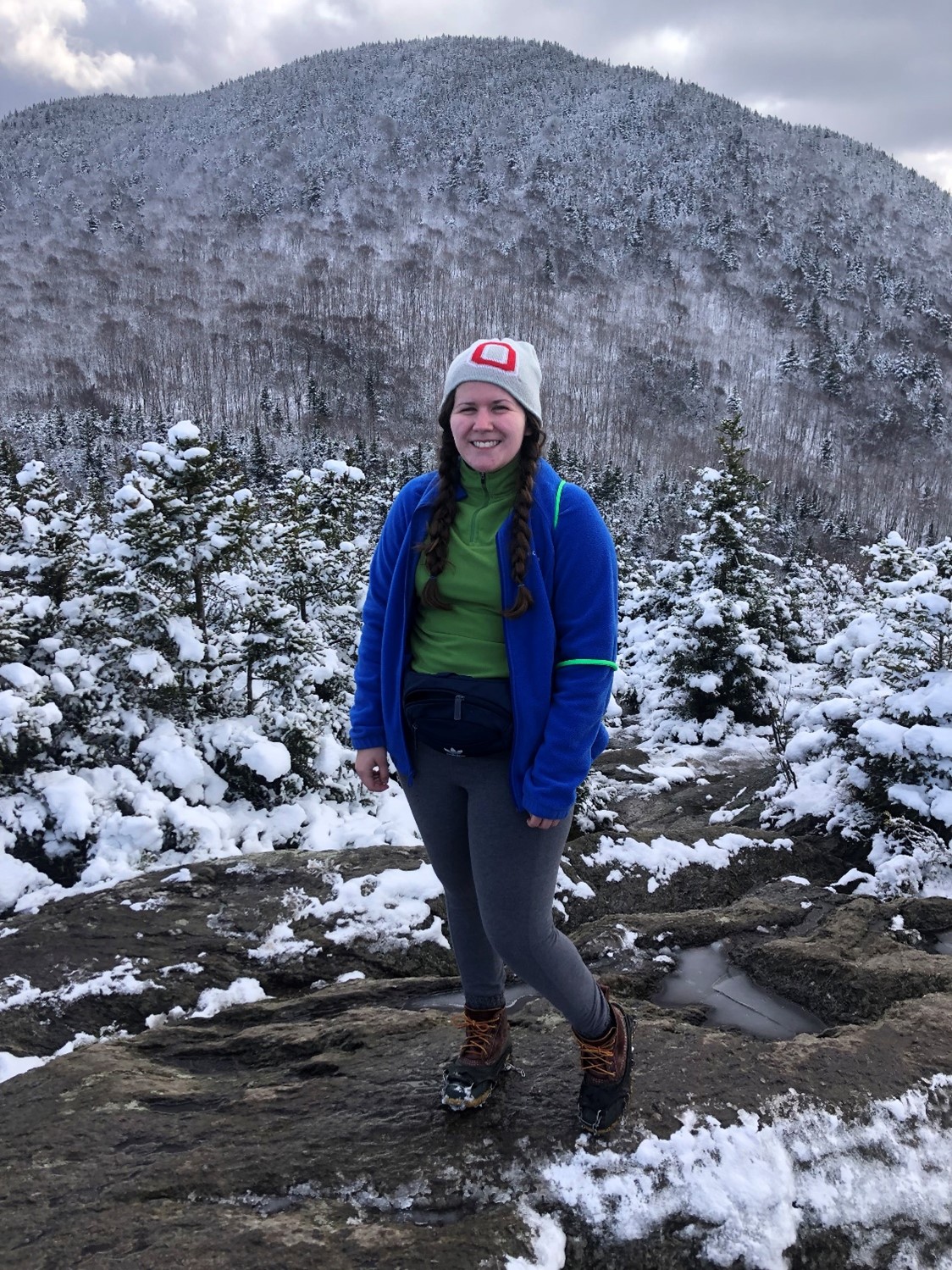 Brittney Welch standing on a muddy trail. Behind her is a large forest and high hill, both covered with a layer of snow