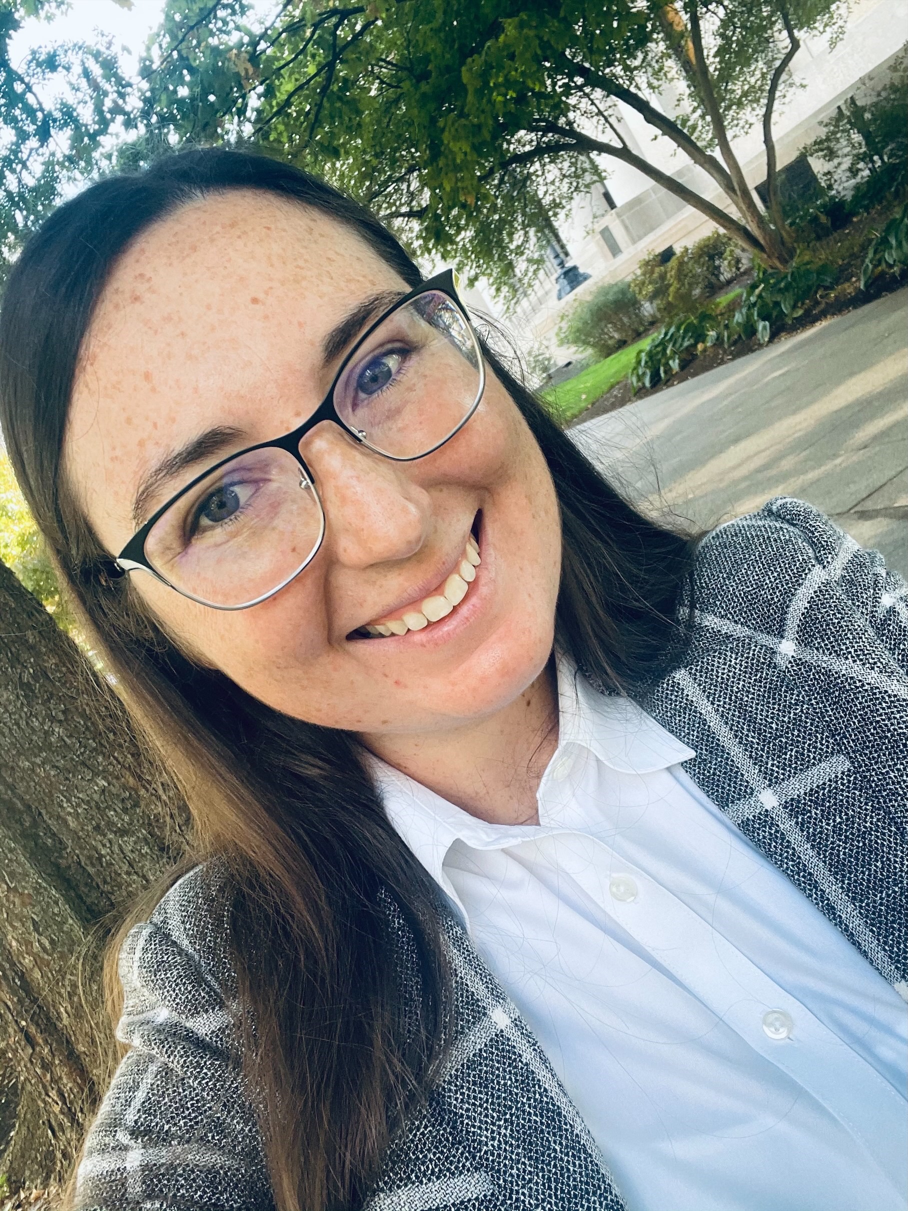 Shea Daley taking a selfie in front of a large building. She is wearing business attire 