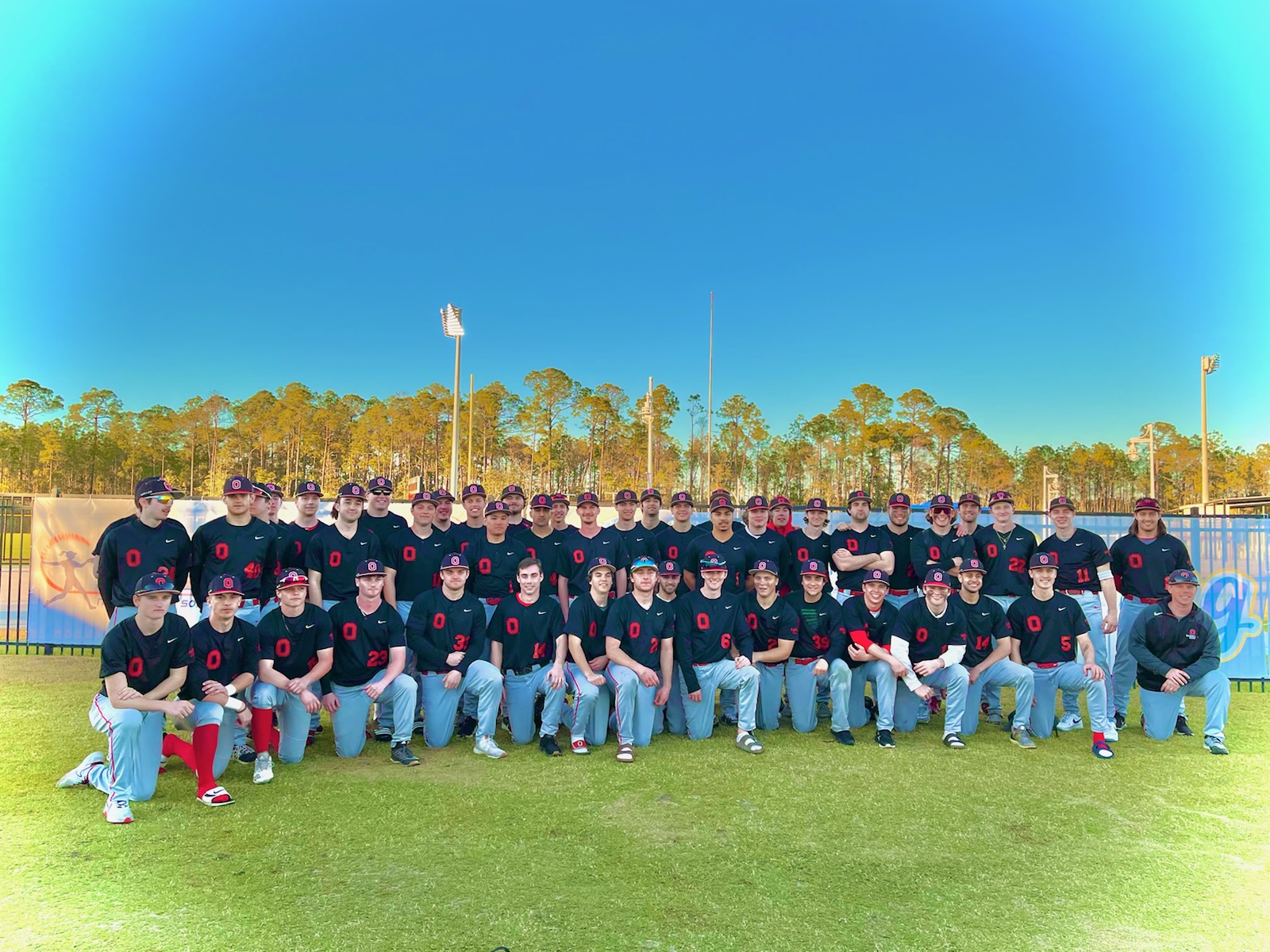 The 2022 Baseball Club in a semi-circle. They pose either standing or on one knee in a baseball outfield