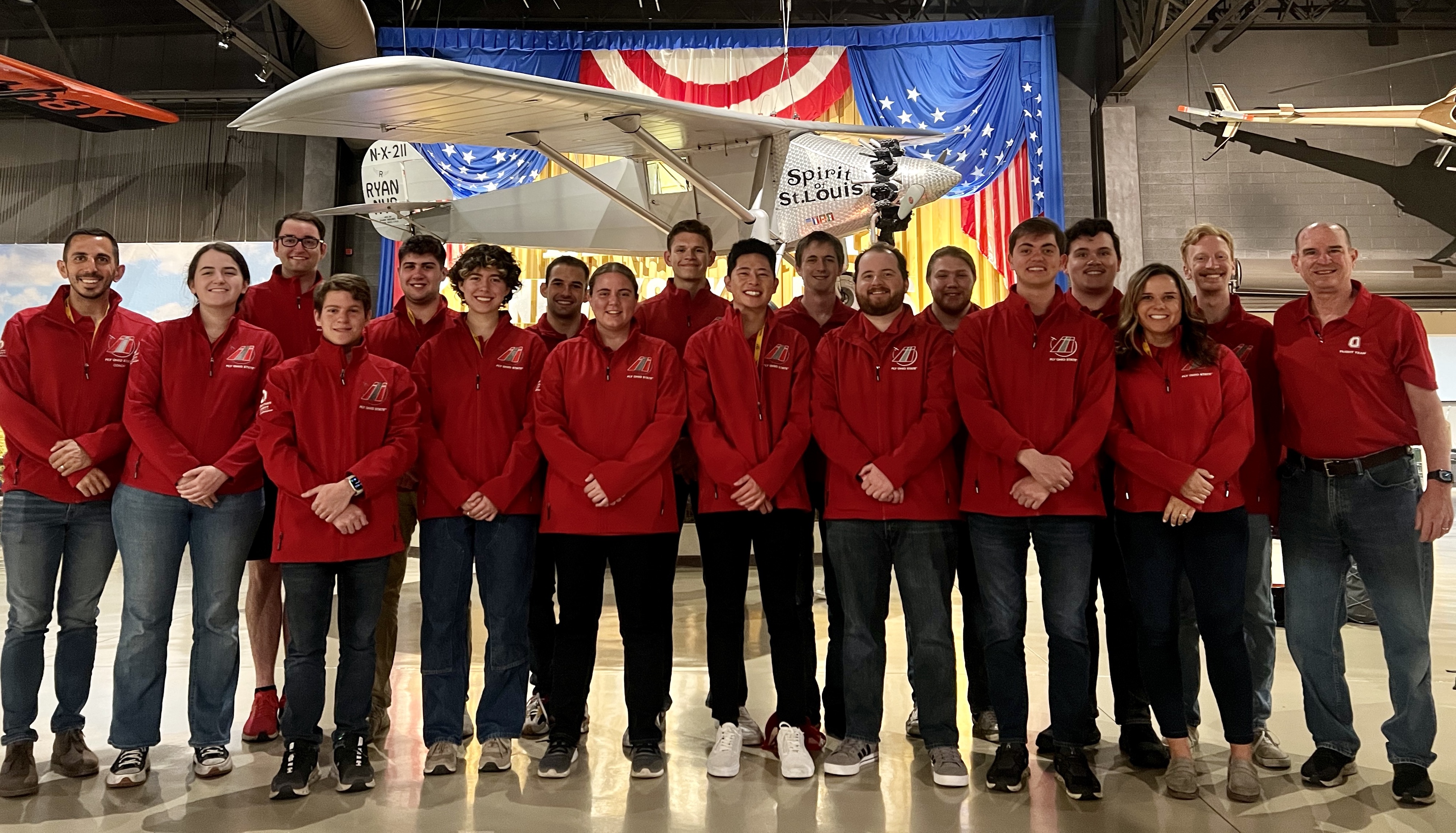 The Ohio State Flight Team posed before a hanging, white plane. They all wear matching half zip jackets
