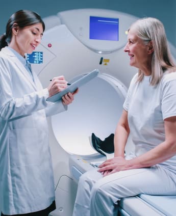 A lady is sitting on the flat bed of a CT scanning machine. She is smiling while speaking to her female radiographer.