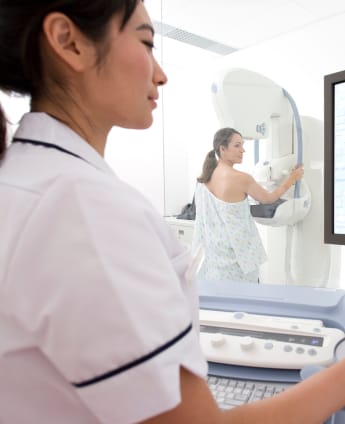 A female mammographer views images on a computer screen while a patient undergoes a mammogram behind a glass window.