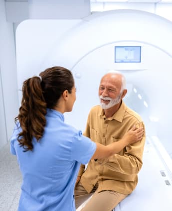 A female radiographer comforts an elderly man who is sitting on the MRI scanner platform before having an MRI scan.