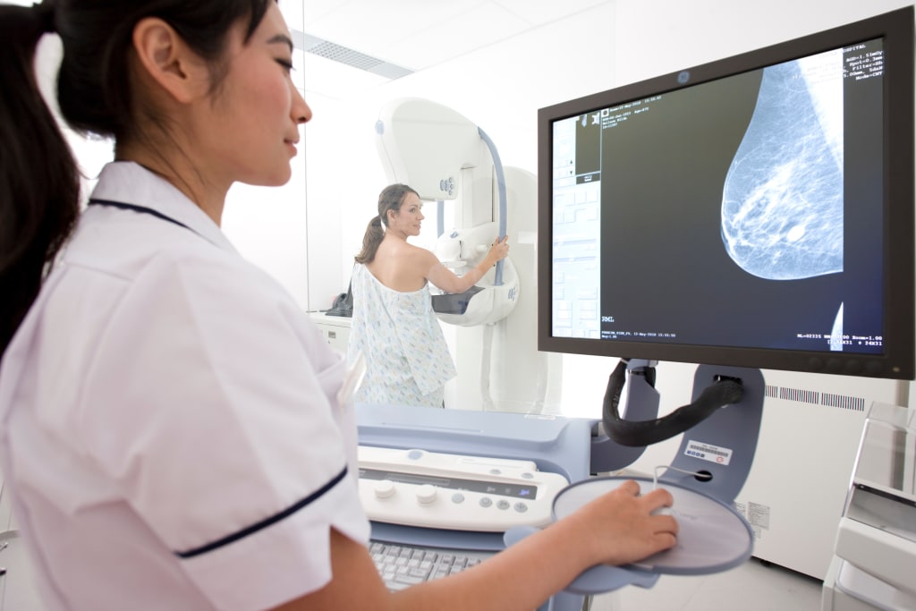 A female mammographer views images on a computer screen while a patient undergoes a mammogram behind a glass window.