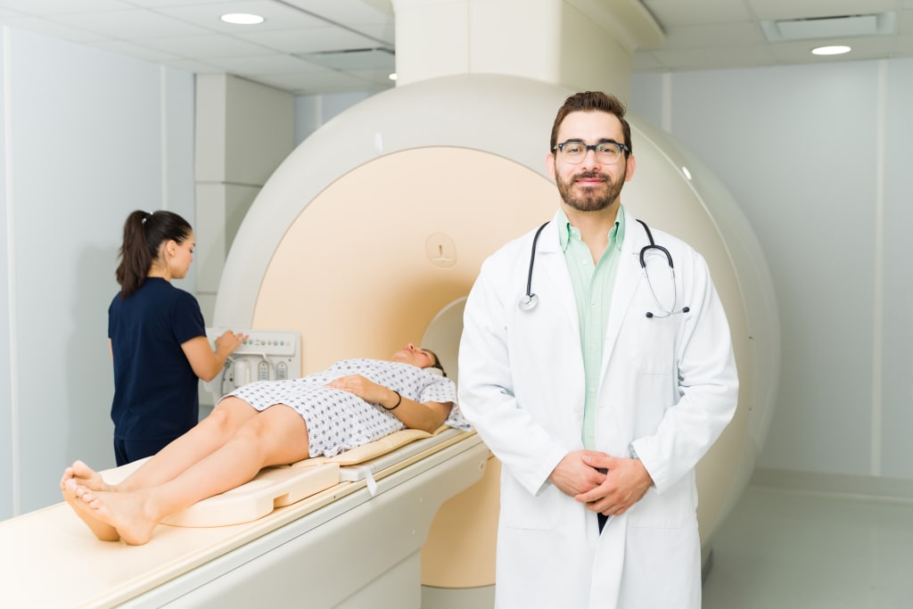 A doctor stands in front of a white MRI scanner while a patient is being prepared for a scan by a radiographer.
