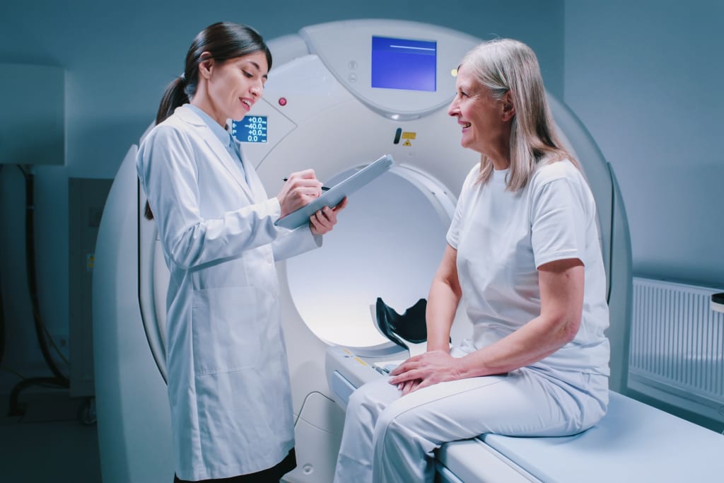 A lady is sitting on the flat bed of a CT scanning machine. She is smiling while speaking to her female radiographer.
