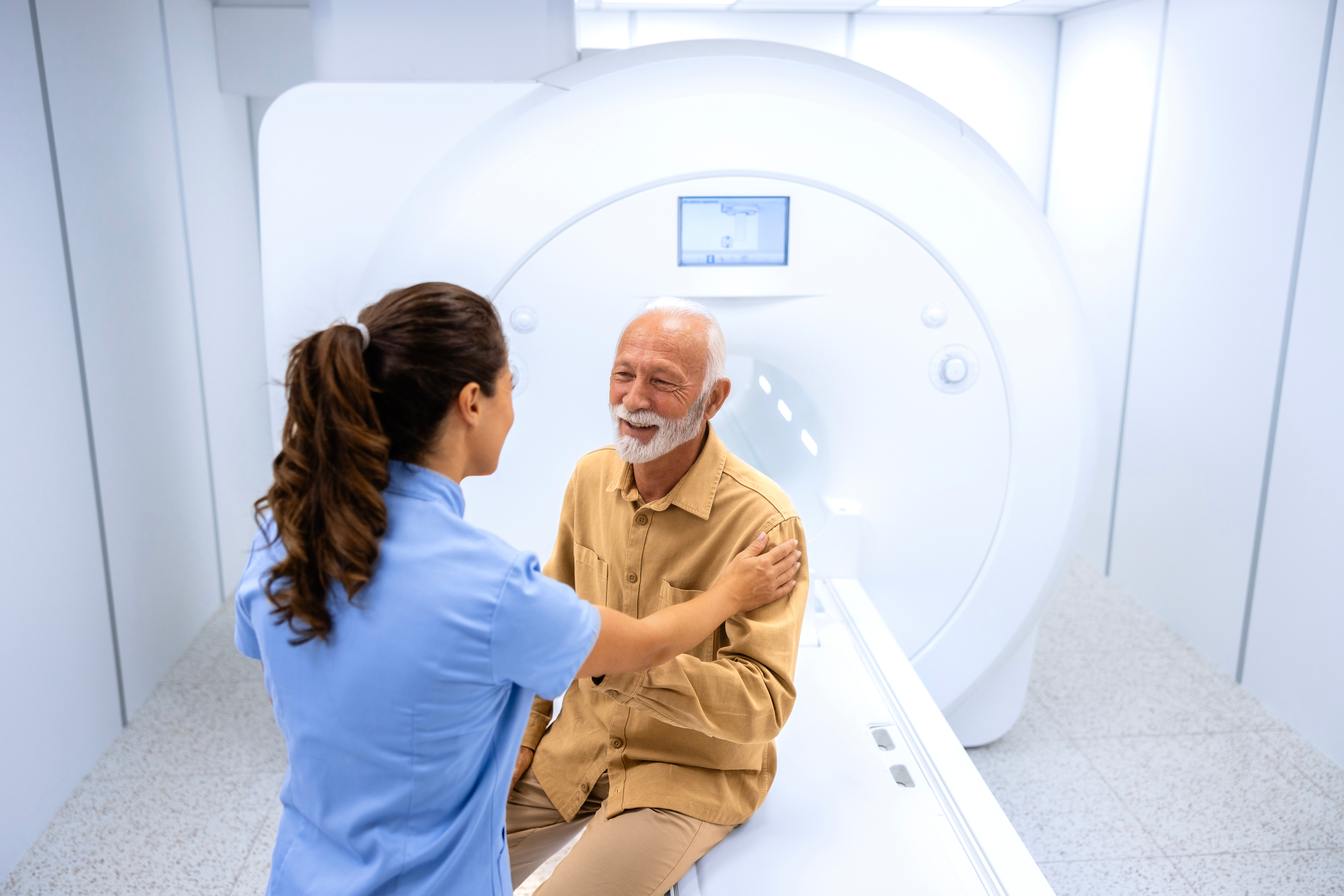 A female radiographer comforts an elderly man who is sitting on the MRI scanner platform before having an MRI scan.
