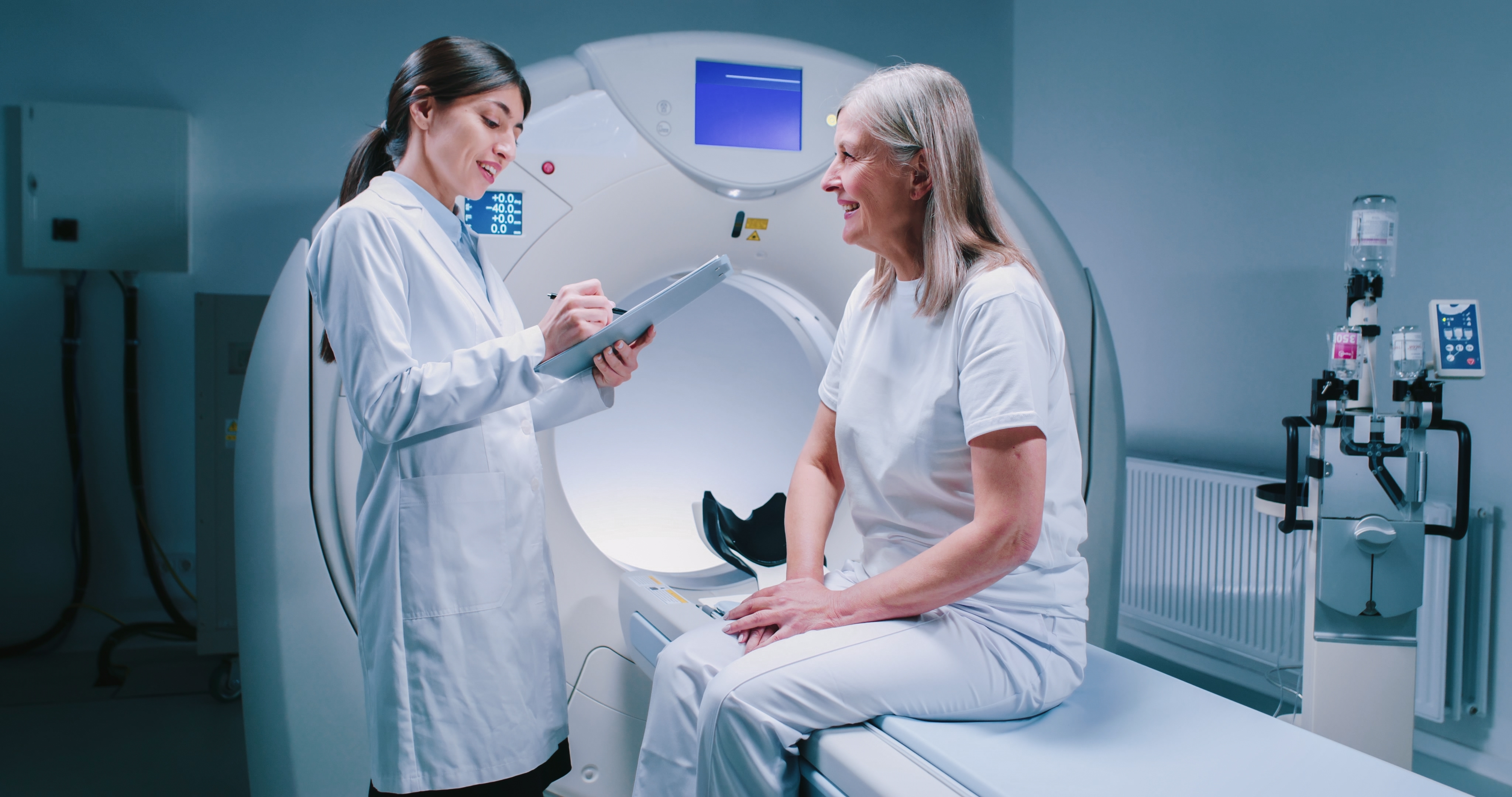 A lady is sitting on the flat bed of a CT scanning machine. She is smiling while speaking to her female radiographer.