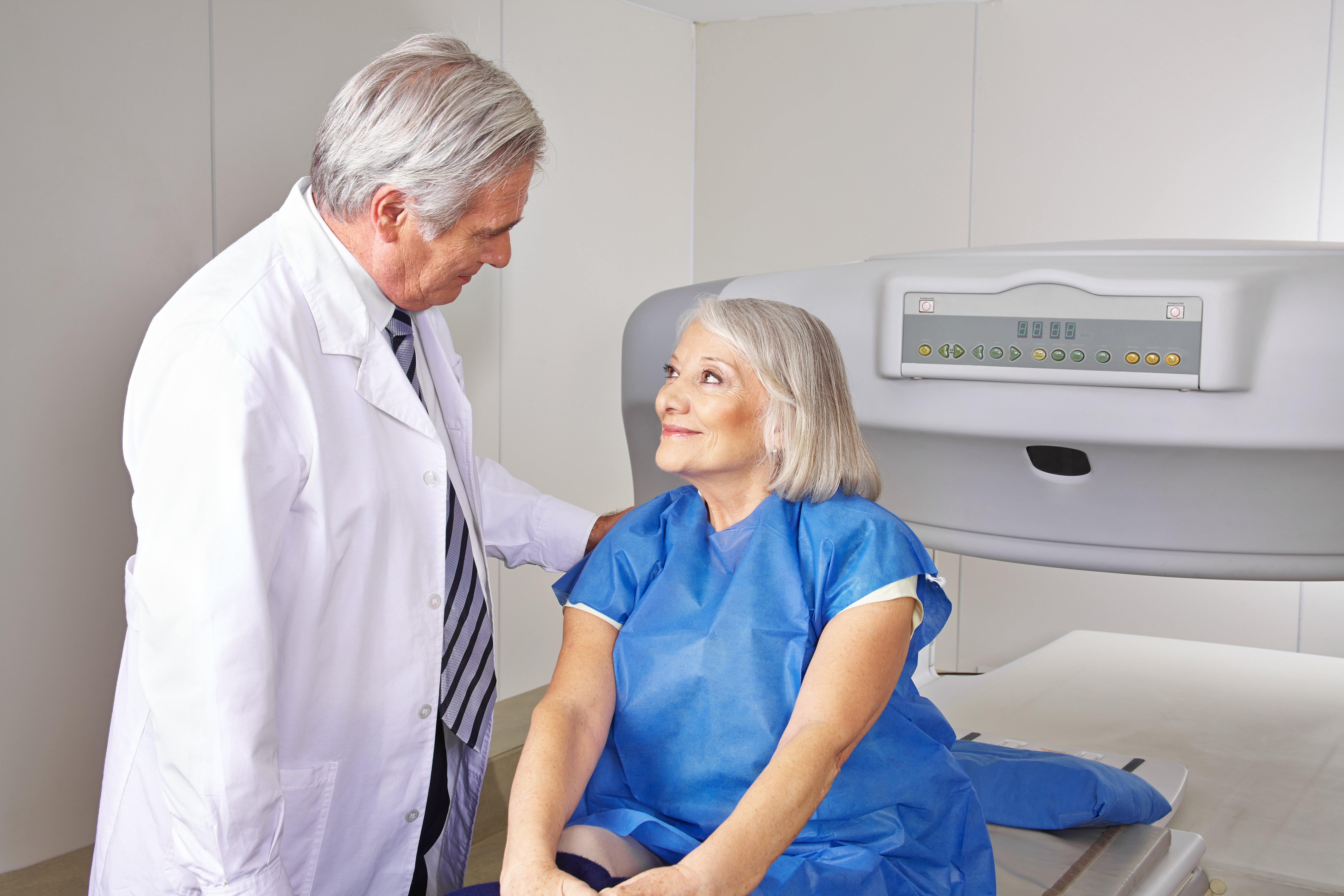 A male doctor in white coat reassures an elderly woman beside a DEXA scanning machine.