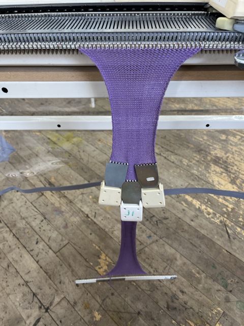 The swatch of purple knitting has become quite long, with the cast-on comb hanging close to the ground. Three claw weights cluster in the middle, providing tension.