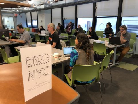 Eleven attendees sit at 4 tables and a back row of chairs. all face the front of the room where the speaker is off-camera