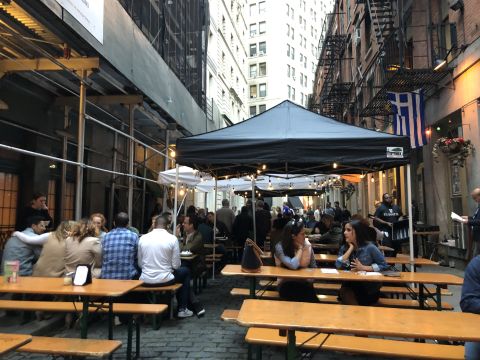 a gray brick street in New York City packed with construction scaffolding and wooden tables, some covered by canopies, a few with people seated at them.