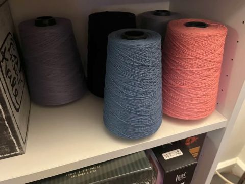 A too-dark photo of yarn cones on a shelf. Blue and pink are visible in front, with purple, black, and gray harder to discern in the back.