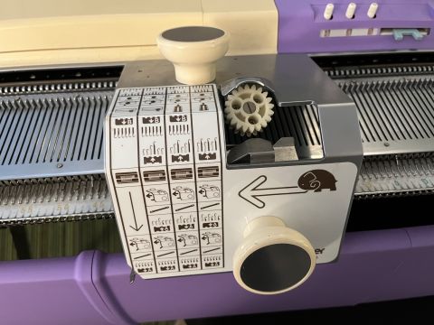 Transfer carriage resting on knitting machine bed. Includes two knobs at 90 degree angles, a visible gear mechanism, and large vinyl decal with instructions featuring an elephant about to charge in the direction the tool is meant to move.