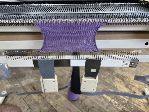 A few dozen rows of plain knitting in purple yarn hanging from the bed. There's evidence on the left of a dropped stitch hastily put back into work.