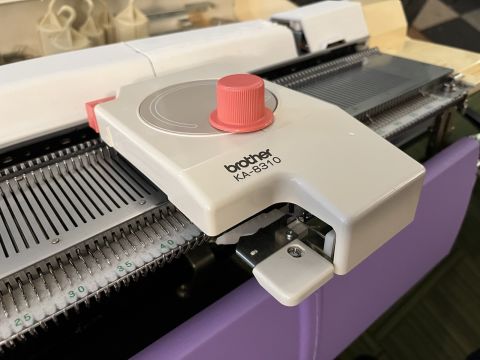 Linker carriage resting on knitting machine bed. Includes bright pink button and knob, as well as an area for stitches to enter.